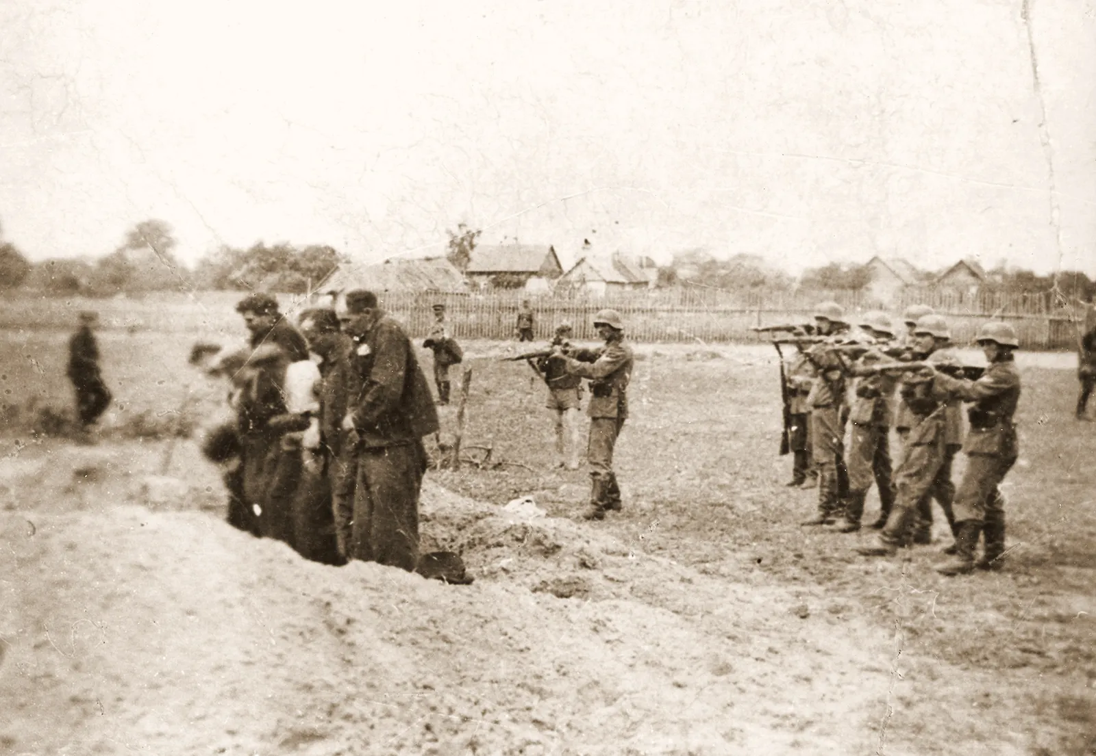 Black-and-white historical photograph showing a group of uniformed soldiers aiming rifles at a line of civilians standing at the edge of a trench in an open field. The civilians appear subdued and closely grouped, while the soldiers stand in formation preparing to fire. In the background, additional figures and small buildings or houses are visible, indicating a rural setting. The image conveys a scene of mass execution during wartime.